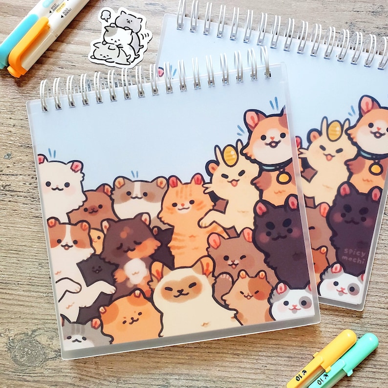 Puede incluir: Una libreta de espiral con una cubierta azul claro que presenta una ilustraci&oacute;n de dibujos animados de un grupo de gatos con diferentes colores de pelaje y expresiones. El texto "Spicy Mochi" est&aacute; impreso en la cubierta.