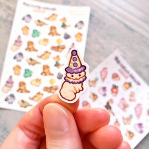 May include: A sticker of a white animal wearing a purple and white party hat. The animal has a purple collar and is smiling. The sticker is on a white background.