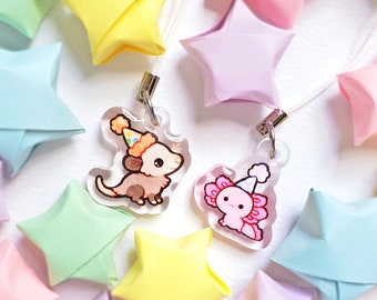 May include: Two colorful charm keychains featuring a pink axolotl and a brown opossum wearing party hats. The charms are attached to white strings and are surrounded by origami stars in various colors.