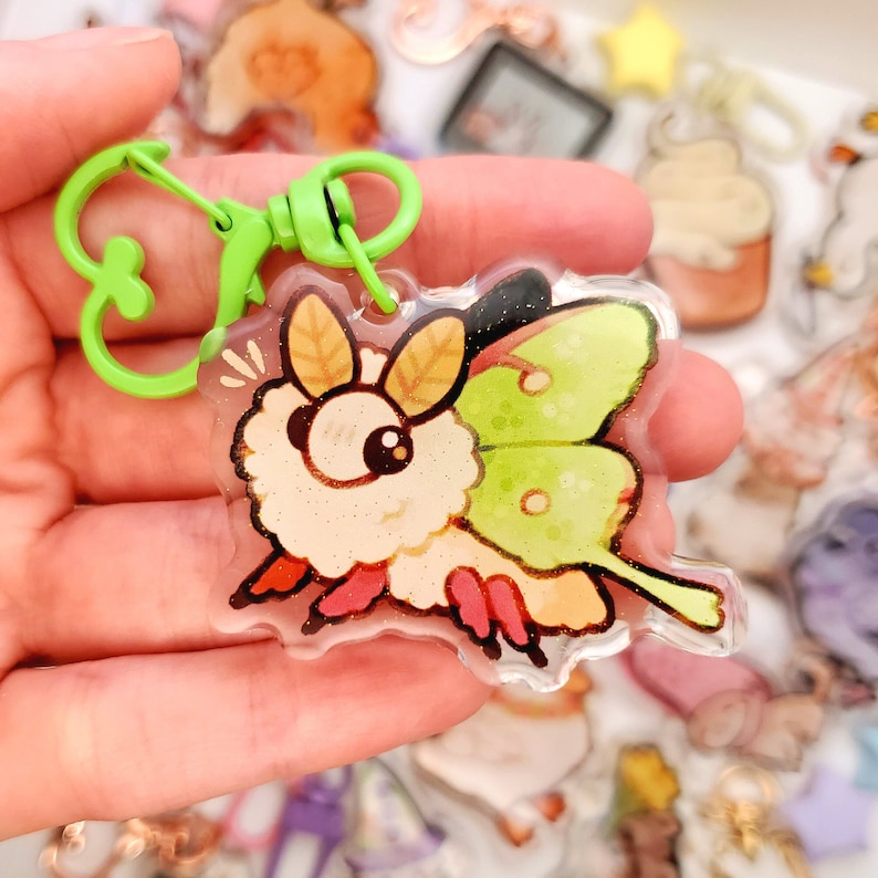 May include: A clear acrylic keychain featuring a cartoon moth with a fuzzy body and green wings. The keychain is attached to a green metal spring clasp.