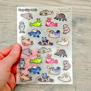 May include: A clear sticker sheet featuring various cartoon seals in different colors and poses. The text "tiny silly seals" is at the top, with the website and social media handle of the seller. The seals have accessories like party hats and bows.
