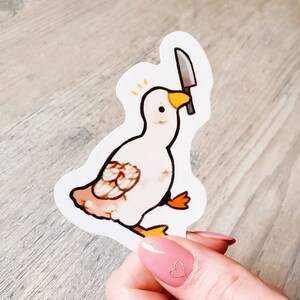 May include: A white goose sticker with a knife in its beak. The goose has orange feet and a yellow beak. The sticker is on a white background.