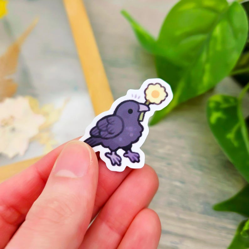 May include: A sticker featuring a cartoon crow holding a yellow flower in its beak. The crow is purple with a white outline, and the flower is yellow with a white center. The sticker is held in a person's hand.