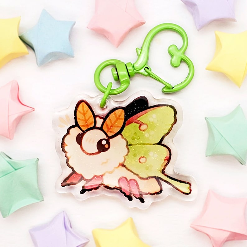 May include: A clear acrylic charm featuring a cartoon illustration of a green and white luna moth with a fluffy white body. The charm is attached to a green metal key chain with a heart-shaped clasp.
