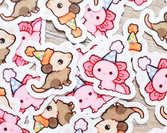May include: A collection of colorful stickers featuring cartoon axolotls and opossums wearing party hats. The stickers are on a light brown wooden surface.