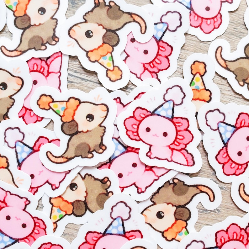 May include: A collection of colorful stickers featuring cartoon axolotls and opossums wearing party hats. The stickers are on a light brown wooden surface.