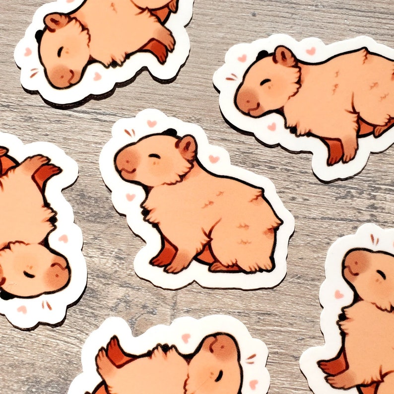 May include: Five stickers featuring cartoon capybaras in various poses. The capybaras are all pink and have black eyes and noses. The stickers are on a light brown wooden surface.