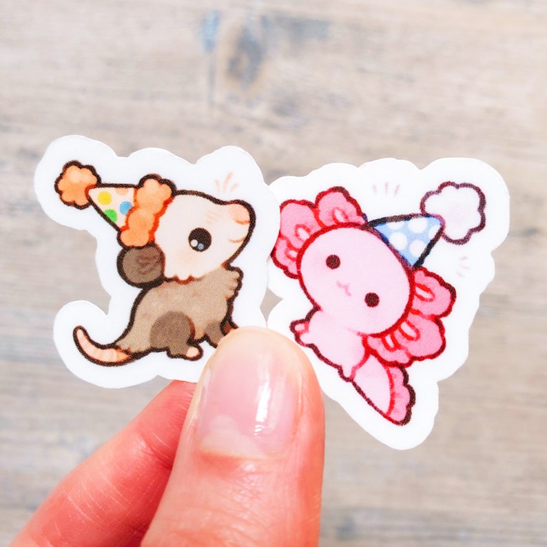 May include: Two adorable animal stickers with party hats. A brown opossum with a yellow and orange hat and a pink axolotl with a blue and white hat.