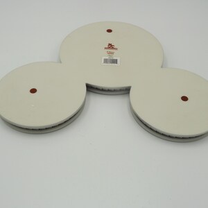 May include: A white ceramic decorative piece with three circular sections connected. Each circle has a dark brown dot in the center. The edges have a textured, dark gray border. A label with text is visible.