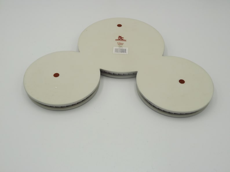 May include: A white ceramic decorative piece with three circular sections connected. Each circle has a dark brown dot in the center. The edges have a textured, dark gray border. A label with text is visible.