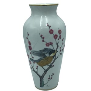 Puede incluir: Jarrón de cerámica blanca con borde dorado, con un diseño pintado de un pájaro posado en una rama floreciente con flores rojas. El jarrón tiene una forma clásica y elegante, adecuado para exhibir flores.