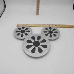 May include: Three silver-colored coasters with a black, floral-like design in the center. The coasters are arranged in a triangular shape. A wooden ruler is placed above the coasters. The coasters are approximately 4 inches in diameter.