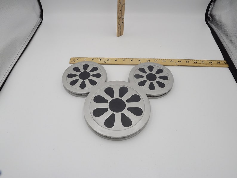 May include: Three silver-colored coasters with a black, floral-like design in the center. The coasters are arranged in a triangular shape. A wooden ruler is placed above the coasters. The coasters are approximately 4 inches in diameter.