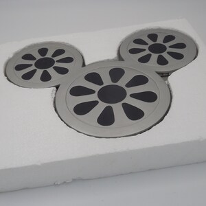 May include: A set of three silver-colored coasters arranged in a Mickey Mouse head shape, each with a black center and a radial design. The coasters are nestled in a white foam insert, suggesting a protective packaging.