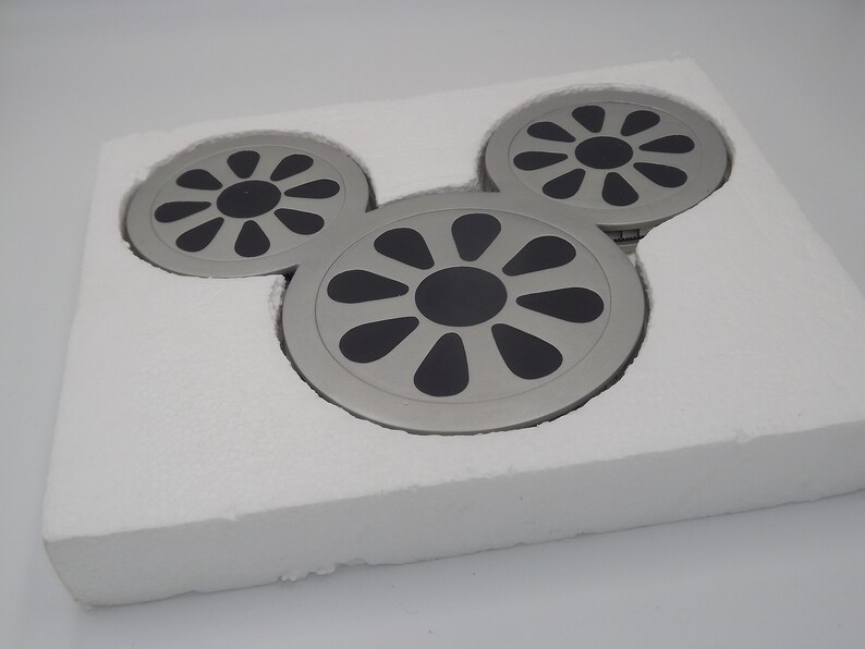 May include: A set of three silver-colored coasters arranged in a Mickey Mouse head shape, each with a black center and a radial design. The coasters are nestled in a white foam insert, suggesting a protective packaging.