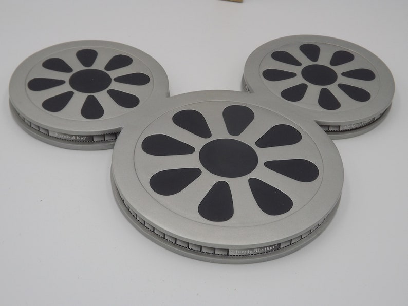 May include: Three gray, circular coasters arranged in a Mickey Mouse head shape. Each coaster has a black, stylized flower design in the center and the text "Musical Kid" and "Lunch Rhythm" on the sides.