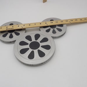 May include: Three silver-colored, circular coasters with black, stylized flower designs. A wooden ruler, marked in inches, rests across the coasters. The coasters are on a white surface.