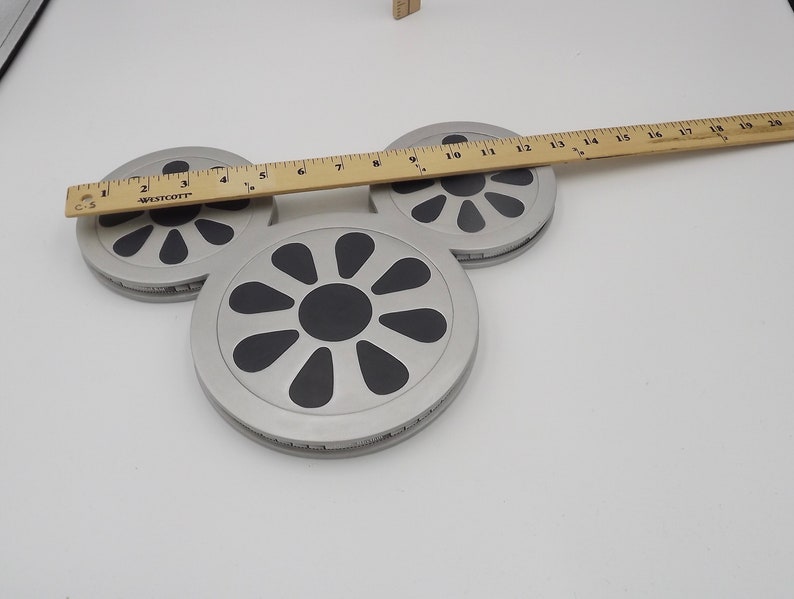 May include: Three silver-colored, circular coasters with black, stylized flower designs. A wooden ruler, marked in inches, rests across the coasters. The coasters are on a white surface.
