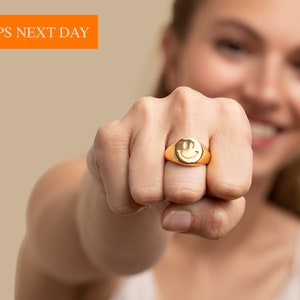 May include: A gold ring with a smiley face design. The ring is worn on the index finger of a person's hand.