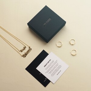 May include: A black box with the text "JWI GANG" on the top, next to a gold necklace with two nameplates that read "Kayla" and "Brianna". There are three gold rings on a white surface. A black and white card with the text "JEWELRY CARE TIPS" is on the white surface.