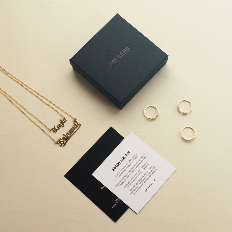 May include: A black box with the text "JWI GANG" on the top, next to a gold necklace with two nameplates that read "Kayla" and "Brianna". There are three gold rings on a white surface. A black and white card with the text "JEWELRY CARE TIPS" is on the white surface.
