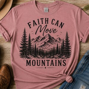 May include: A dusty rose short-sleeved t-shirt featuring a black graphic print of a mountain range and pine forest. The text "FAITH CAN MOVE MOUNTAINS" is printed in black lettering above and below the landscape illustration on the front.
