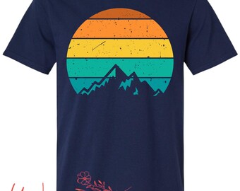 Puede incluir: Camiseta azul marino con un diseño retro de puesta de sol con rayas naranjas, amarillas y turquesas sobre una silueta de montaña. La camiseta tiene cuello redondo y mangas cortas. El logotipo de Wildflowers está en la parte inferior.