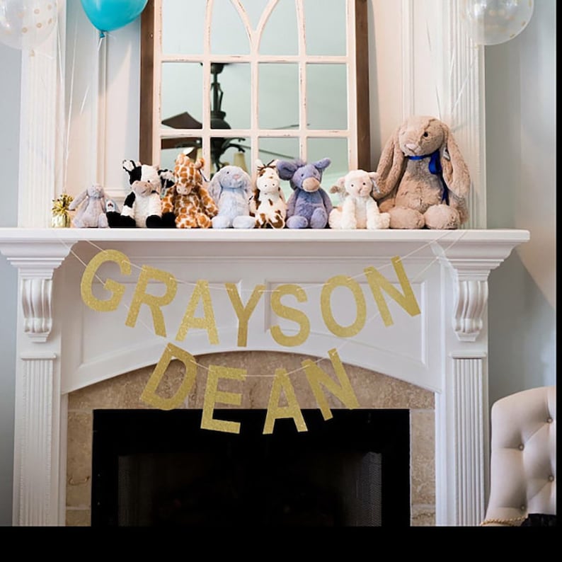 May include: A collection of plush animal toys, including a giraffe, cow, and elephant, are arranged on a white mantel. A gold glitter banner spells out "GRAYSON DEAN". Balloons and a mirror are in the background.