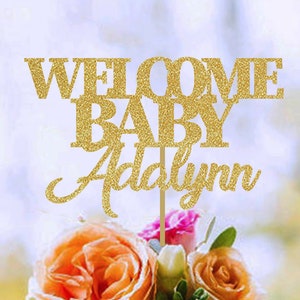 May include: A gold glitter cake topper with the words "WELCOME BABY Addalynn" on a wooden stick. The topper is placed in a bouquet of orange, pink, and white flowers.
