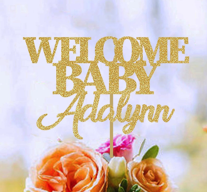 May include: A gold glitter cake topper with the words "WELCOME BABY Addalynn" on a wooden stick. The topper is placed in a bouquet of orange, pink, and white flowers.