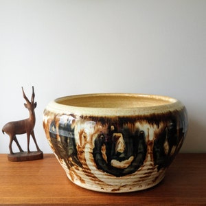 May include: A large, round ceramic bowl with a wide opening. The bowl has a cream-coloured interior and rim, with a dark brown, swirling pattern on the exterior. A small wooden deer figurine stands to the left of the bowl.