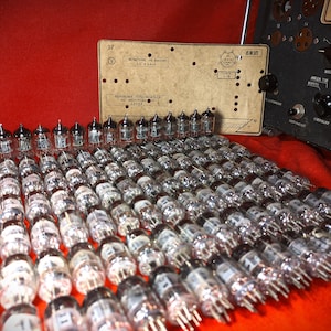 May include: A large collection of vintage vacuum tubes, arranged in rows on a red surface. The tubes are made of glass and metal, and have a variety of shapes and sizes. A printed circuit board and a vintage electronic device are visible in the background.