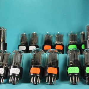 May include: A collection of vintage vacuum tubes with clear glass bulbs and black bases. Some tubes have labels with numbers and letters. The tubes are arranged on a light blue surface, suggesting a focus on electronics or vintage audio equipment.