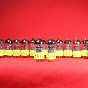 May include: A row of 16 vintage vacuum tubes, each with a silver metal casing and a clear glass section. The tubes are arranged in a line on a red surface. Each tube has a yellow sticker with handwritten numbers on it.