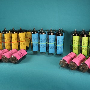 May include: A collection of vintage vacuum tubes with yellow, blue, and pink sticky notes attached. The notes have handwritten text on them, including the numbers "1-48", "1-49", "1-50", and "S-104".