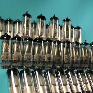 Puede incluir: Una colección de tubos de vacío vintage, dispuestos en forma de pirámide, sobre un fondo azul turquesa. Los tubos son de vidrio y metal, con una parte superior negra y una base plateada.