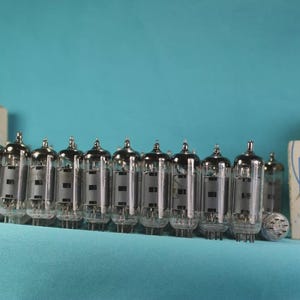 May include: A collection of vintage vacuum tubes with silver tops and glass bodies, arranged in a row. The tubes are accompanied by several small, rectangular boxes with blue and white graphics and Japanese text. The background is a solid teal color.