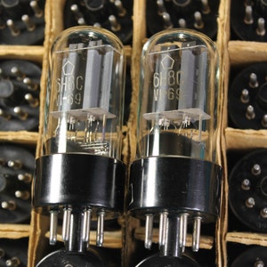 Puede incluir: Dos tubos de vacío vintage, etiquetados como "6H8C VI-69", en una caja de cartón con otros tubos. Los tubos son de vidrio transparente con bases de metal negro y pines plateados.