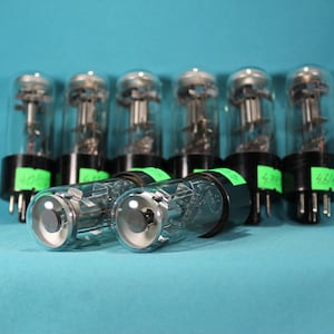 May include: Eight vintage vacuum tubes with black bases and silver tops. The tubes are arranged in a row on a blue surface. The tubes have green labels with numbers on them.