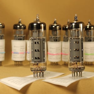 May include: A collection of vintage vacuum tubes with clear glass bulbs and metallic tops. Each tube has a white label and is secured with a colored rubber band. The tubes are arranged on a light brown surface.