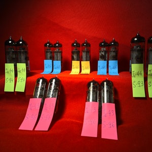 May include: A collection of vintage vacuum tubes with handwritten labels on yellow, green, blue, and pink sticky notes. The labels include numbers and letters, such as "J-44 S-94" and "1-43 S-96".