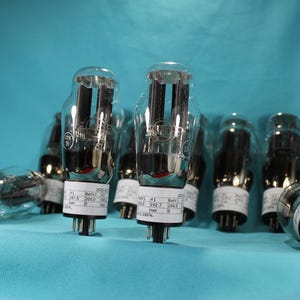 May include: A group of eight clear glass vacuum tubes with metal caps and labels. The tubes are arranged in a row on a blue surface. The labels on the tubes include text such as "200.0", "100.0", "180.0", and "100.0".