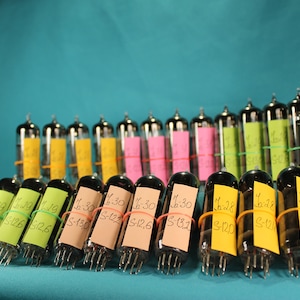 May include: A collection of vintage vacuum tubes with handwritten labels, including numbers like "6-30" and "S-120", are arranged in rows on a turquoise surface. The tubes are secured with rubber bands and have a clear glass exterior.