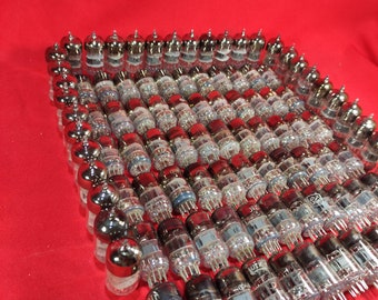 May include: A large collection of vintage vacuum tubes, arranged in a grid pattern on a red surface. The tubes are made of glass and metal, and have a silver finish.