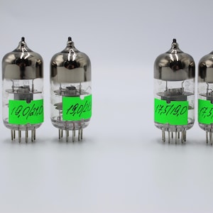 Op de afbeelding: Vier vintage vacuümbuizen met zilveren toppen en heldere glazen lichamen. Elke buis heeft een groen label met handgeschreven nummers. De buizen hebben meerdere pinnen aan de basis en zijn gerangschikt op een wit oppervlak.