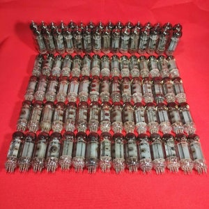 May include: A large group of vintage vacuum tubes, arranged in rows on a red surface. The tubes are made of glass and metal, and have a black and silver finish.