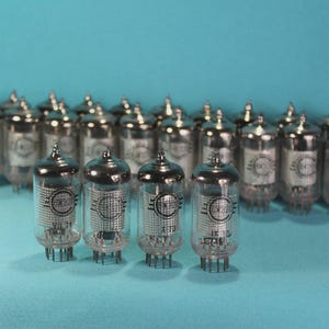 May include: A collection of vintage vacuum tubes with silver tops and clear glass bodies. The tubes are arranged in rows against a teal background. Each tube has a metal base with pins and internal components visible through the glass.