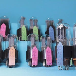 Puede incluir: Una colección de válvulas de vacío antiguas con bombillas de vidrio transparente y bases negras. Cada válvula tiene una etiqueta de color con números escritos a mano. El fondo es azul claro liso.