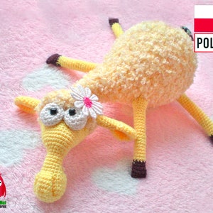 Pode incluir: Um brinquedo de cordeiro amarelo de crochê com pés marrons e uma flor branca na cabeça. O cordeiro está deitado em uma superfície rosa felpuda. O texto "POLSKI" é visível no canto superior direito da imagem.