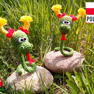 May include: Two handmade crochet snakes with green bodies, yellow and red accents, and googly eyes rest on a rock in a grassy area. The snakes have red tongues sticking out. The image includes the text "POLSKI".